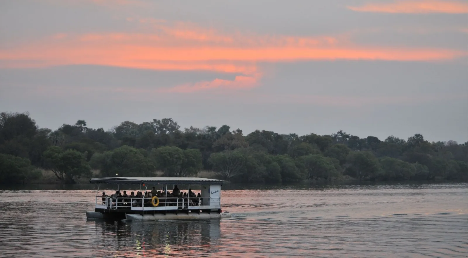 Zambezi River cruise boat at dusk with passengers on the open deck, calm water reflecting a pink and grey twilight sky
