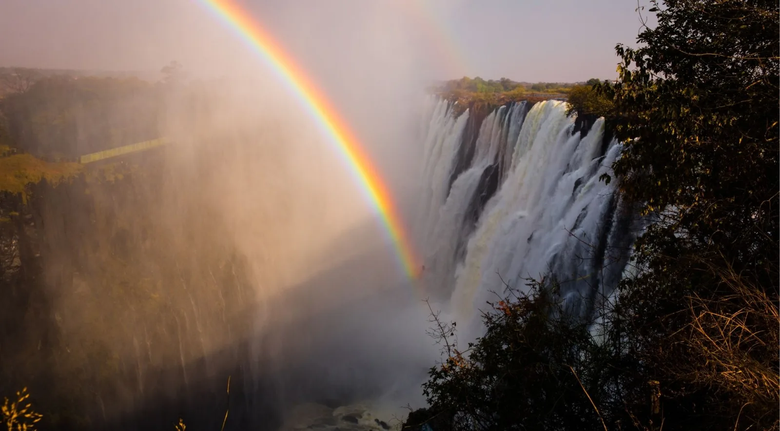 Victoria Falls with a vivid rainbow arching through the rising mist and spray, golden light at the gorge rim