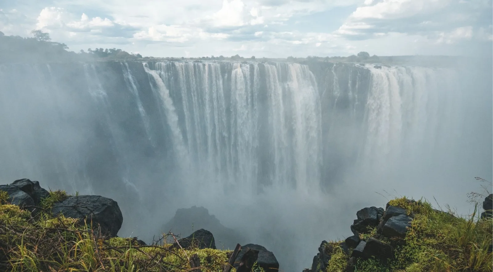 Panoramic front-on view of Victoria Falls — a massive curtain of white water plunging into the gorge with mist rising into a cloudy sky