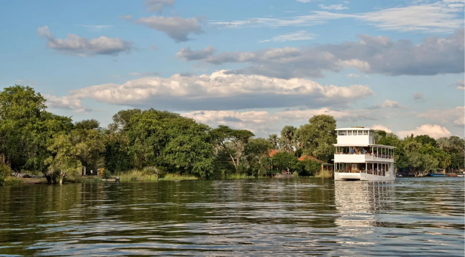 White multi-deck Zambezi River cruise boat on a wide stretch of the river, green riverine trees on both banks under a blue sky